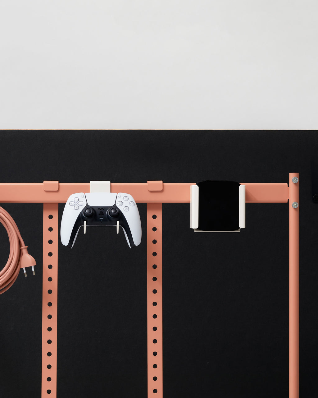 Dusty Rose Apple TV Mount holding a black streaming box on a pink wall rail beside a PS5 controller and cable