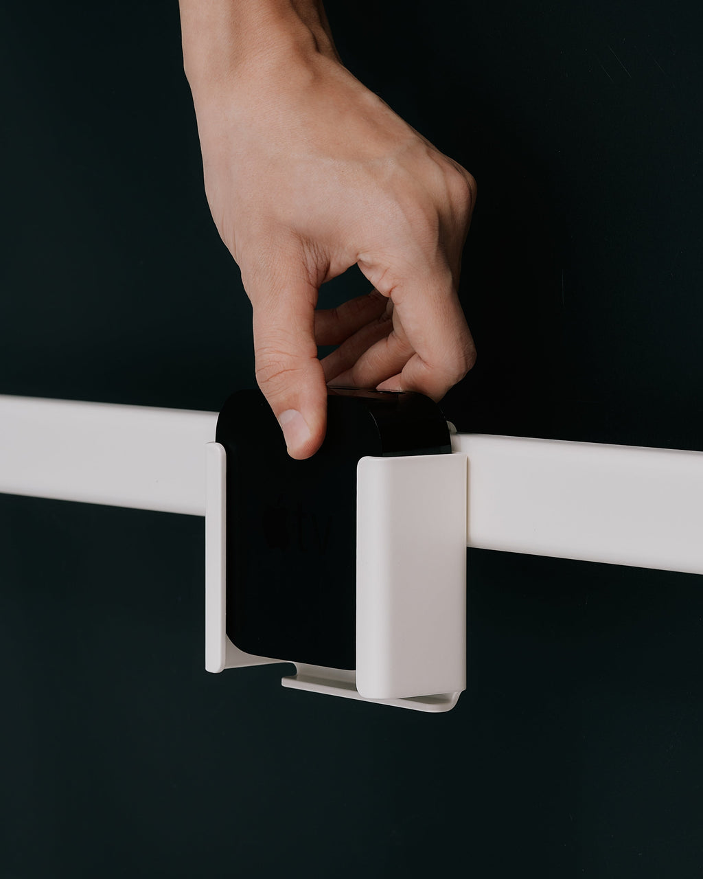 Hand placing an Apple TV into a Bubble Gum white wall mount bracket on a rail against a dark studio backdrop