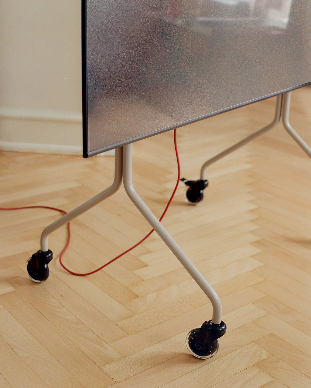 Black Blade Wheels on a beige TV stand's angled legs, shown in a herringbone parquet room with red cable