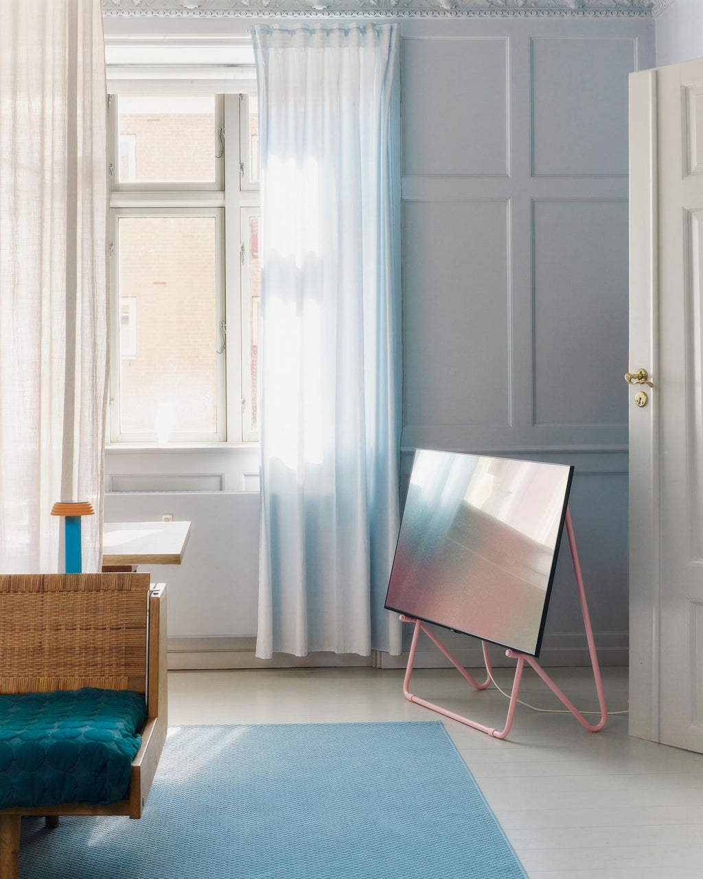 Bloom in Bubble Gum low stand holding a TV in a pastel blue living room, angled view beside sheer curtains