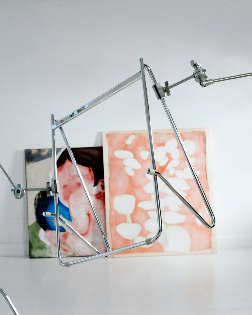 Bloom in Chrome (Stands Low) angled in a bright studio, chrome tubular frame set against leaning artwork panels