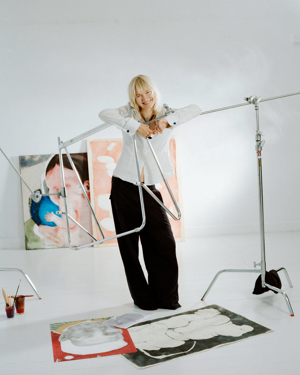 Smiling artist leaning on Bloom in Chrome (Stands Low) frame in a white studio with paintings and light stands
