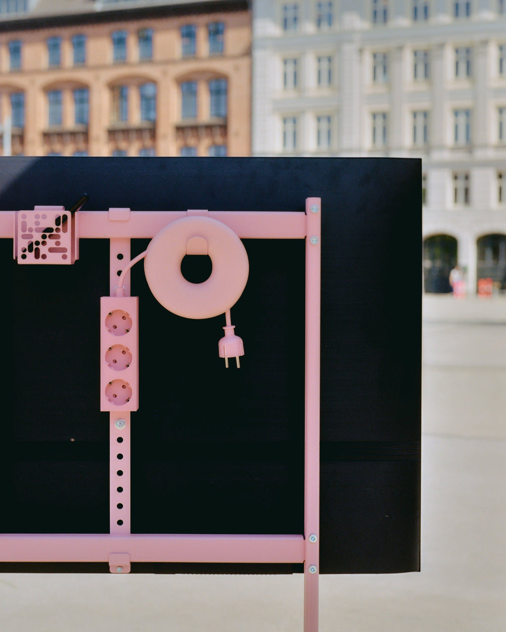 Bubble Gum Cable Bagel winding a pink cord on a black TV back frame outdoors, plug dangling, city buildings behind