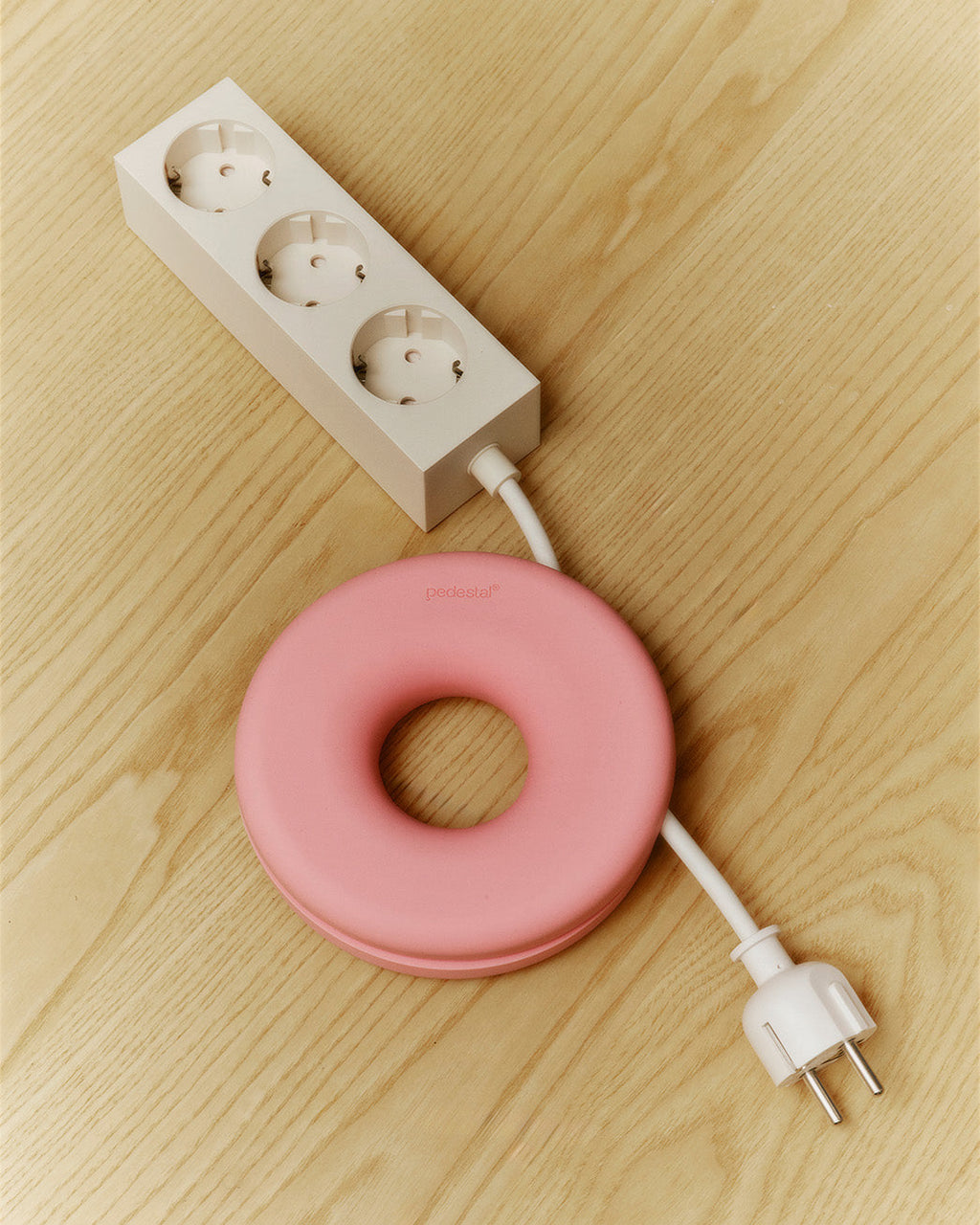 Bubble Gum pink Cable Bagel cable manager on light wood floor, winding a white power strip cord toward its plug