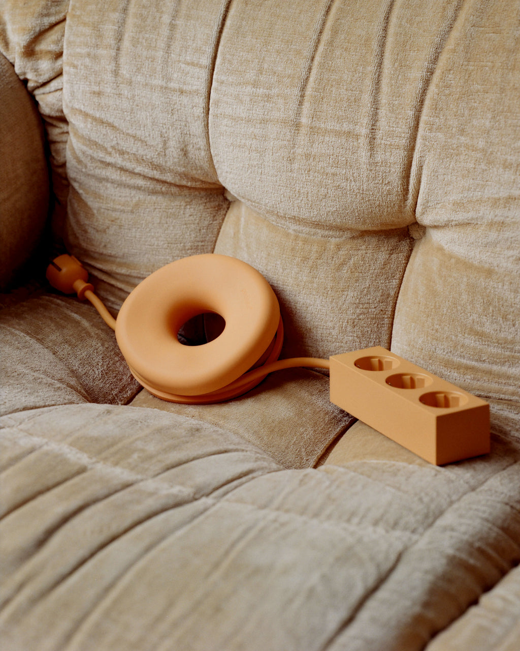 Apricot Cable Bagel coiling a matching power strip cord on a beige chenille sofa, angled lifestyle view