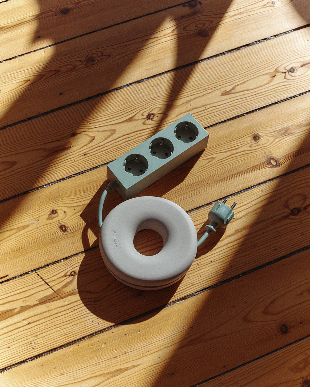 Oat Cable Bagel coiling a sage extension cord on sunlit pine floorboards, beside a matching power strip