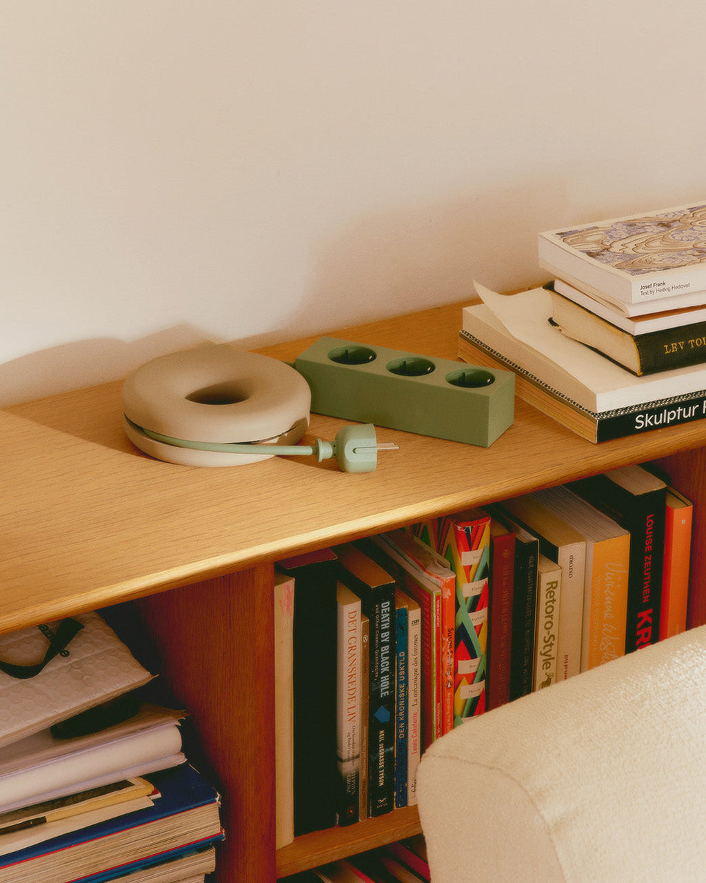 Oat Cable Bagel winding a green cord on a wooden shelf beside a power strip and stacked books in a cozy room