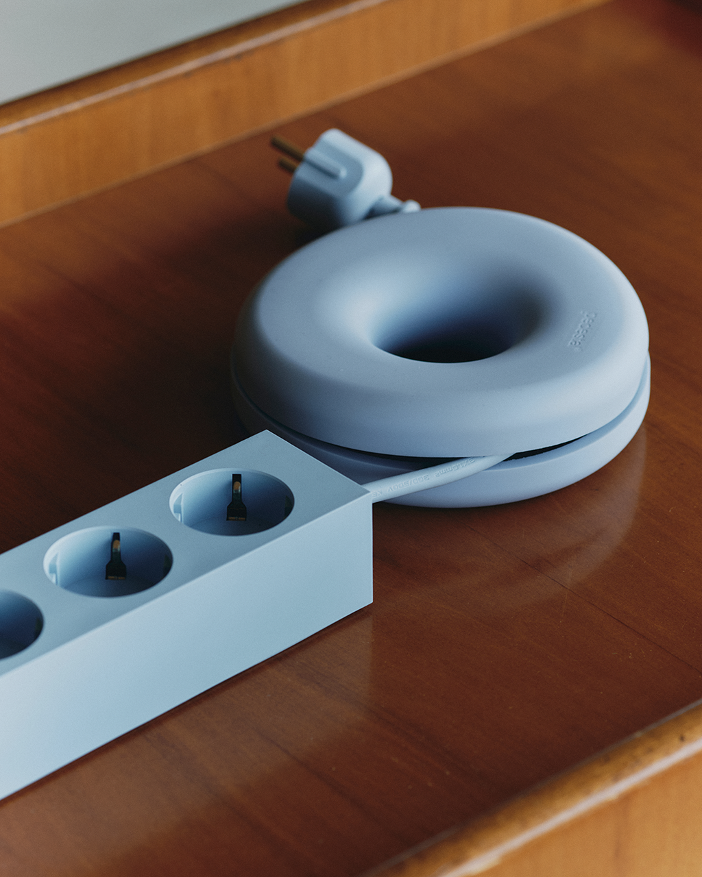 Powder Blue Cable Bagel on a wood desk, winding a matching power strip's cable, seen from a close overhead angle