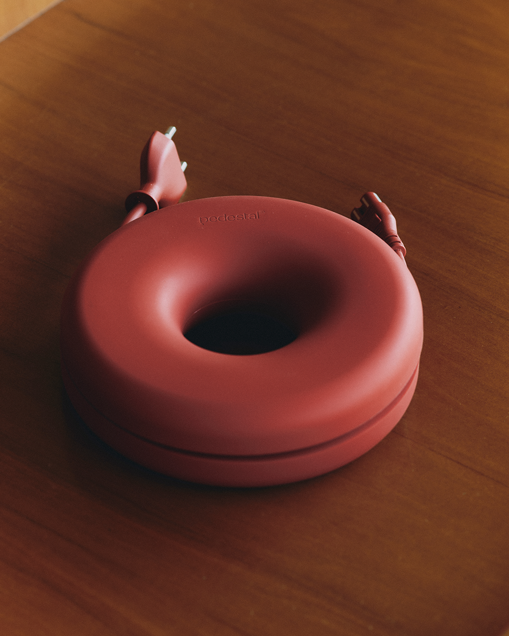Cable Bagel in Burnin' Red resting on a wood table, cord coiled inside with two plug ends emerging at the top