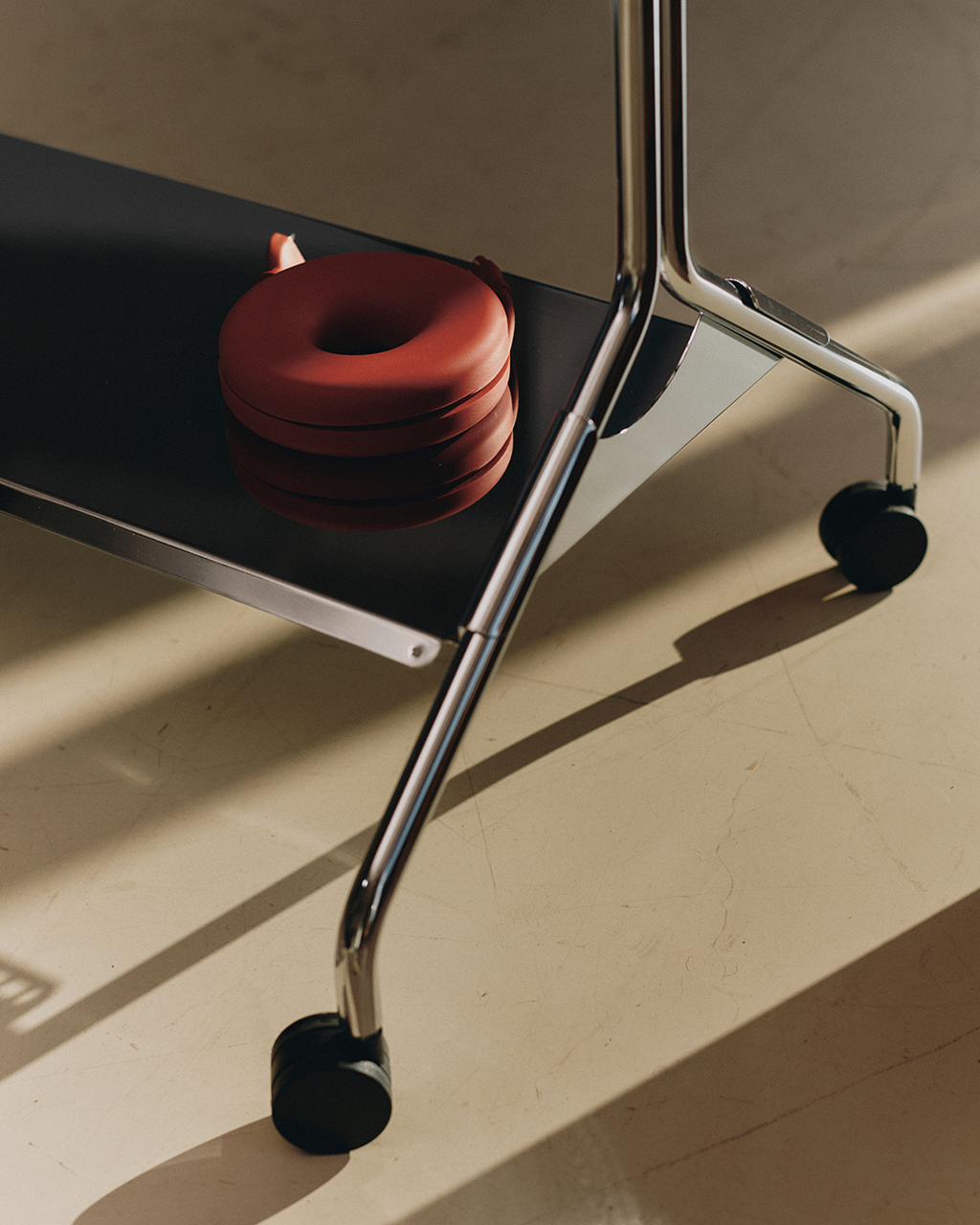 Cable Bagel in Burnin' Red coiled on a black desk shelf, overhead view with chrome legs and casters in sunlight