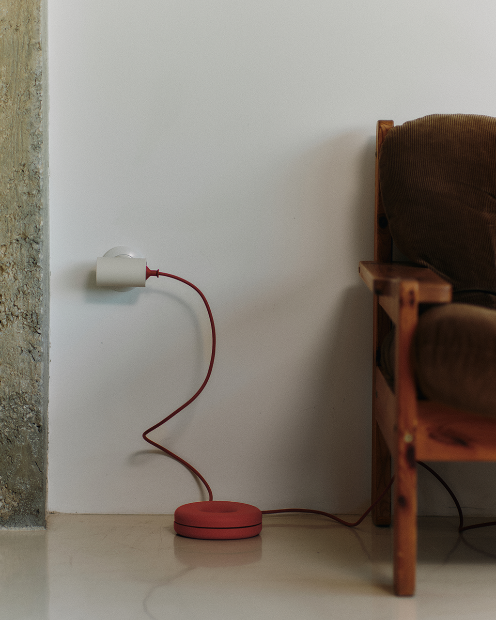 Burnin' Red Cable Bagel on a pale floor coiling a red lamp cord, beside a wooden armchair in a sunlit corner