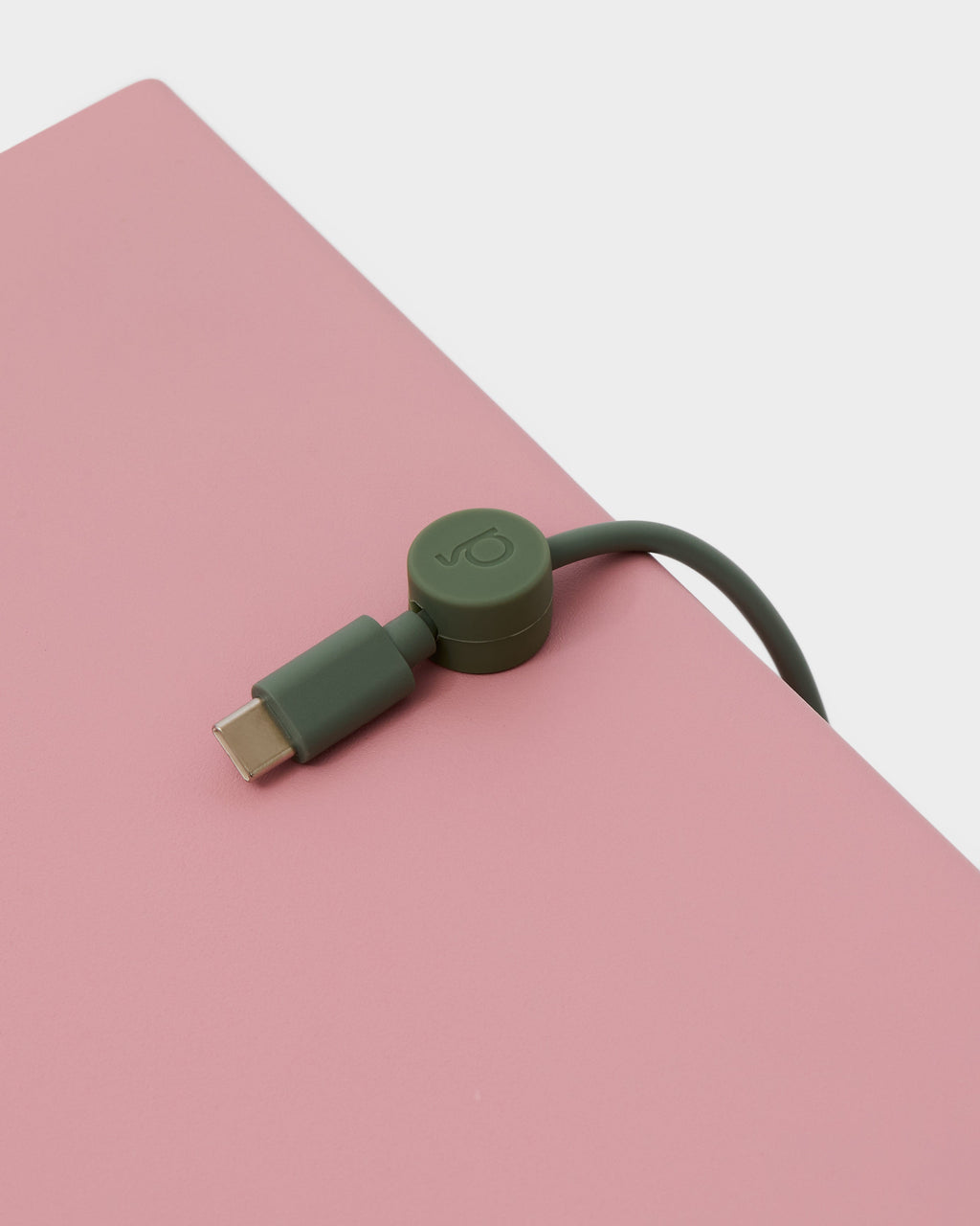 Mossy Green Cable Clip gripping a USB-C cable at the edge of a pink desk, close-up studio detail shot