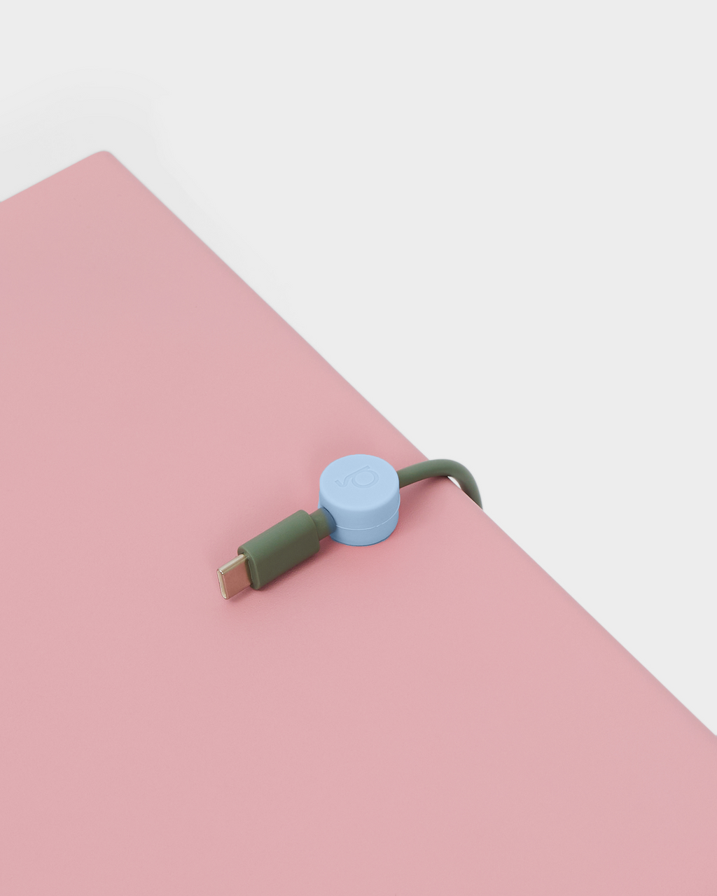 Powder Blue cable clip holding an olive USB-C cable along the edge of a pink desk pad, angled overhead studio view