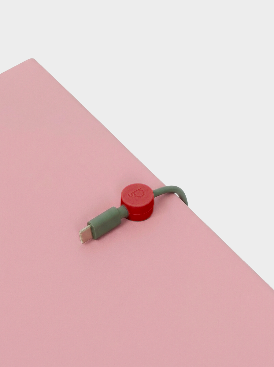 Burnin' Red cable clip gripping a sage green USB-C cable on a pink surface, close-up angled studio detail shot