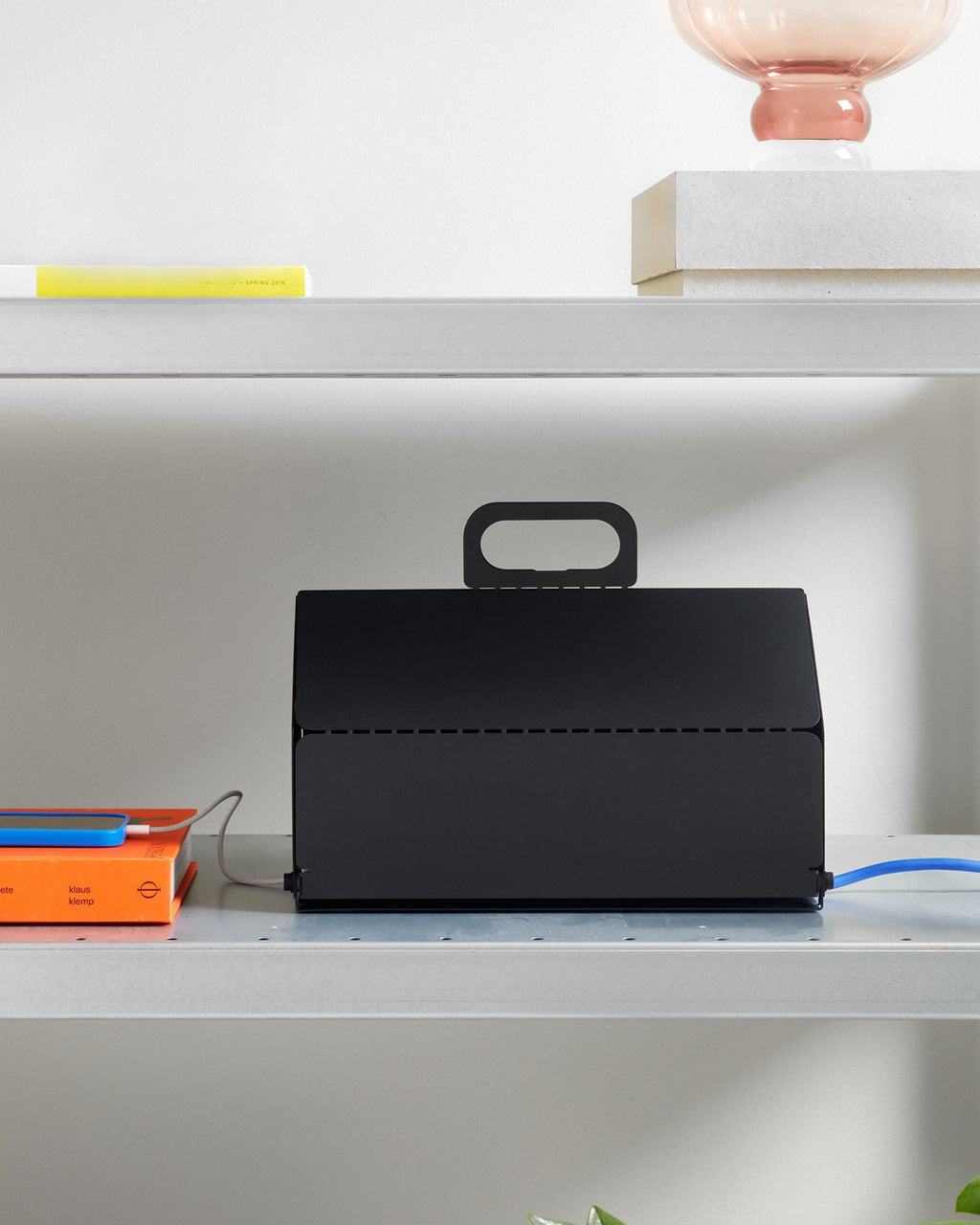 Charcoal Cable House on a metal shelf hiding cords, with blue cable exiting the side beside books and a phone