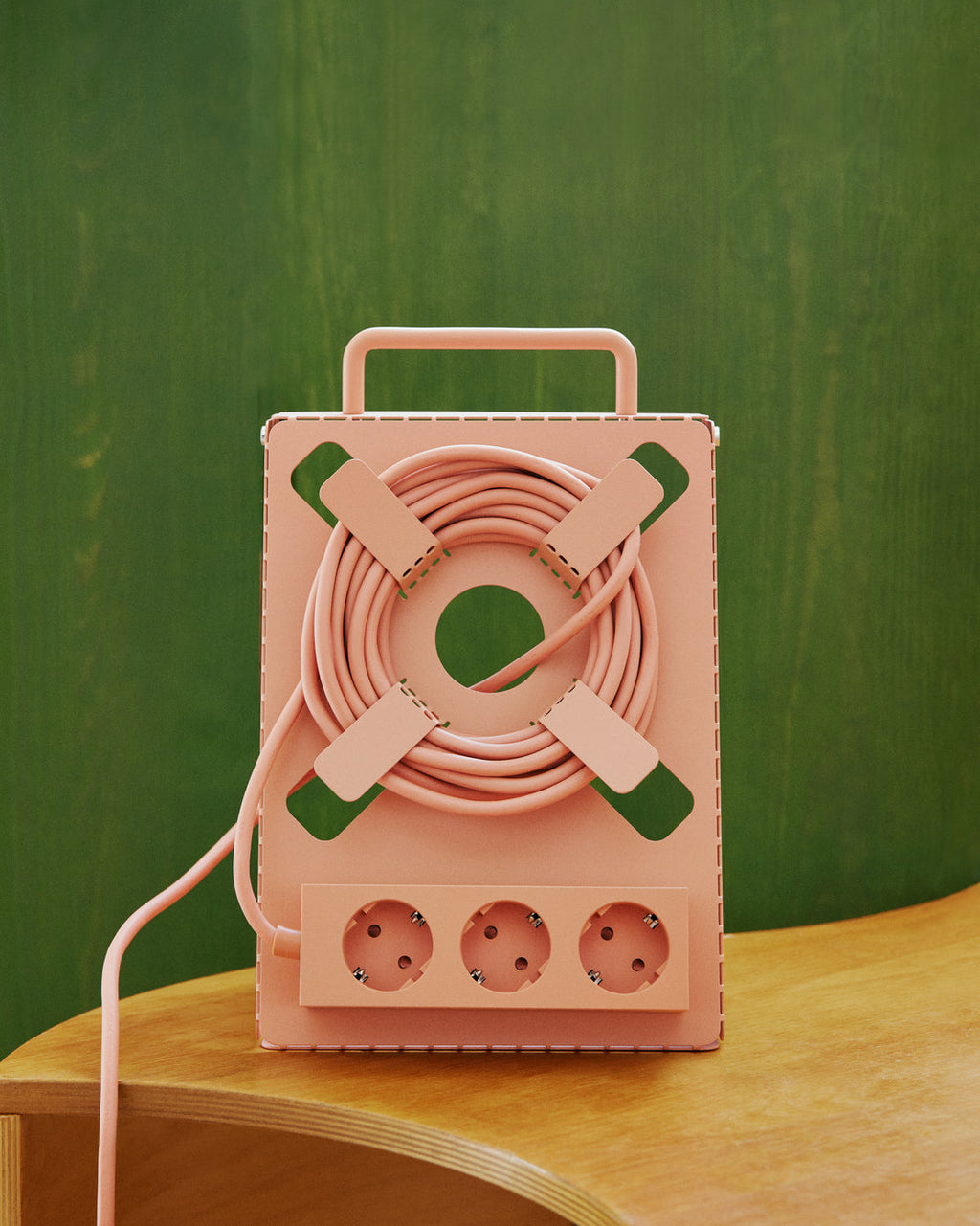 Dusty Rose Cable Stand on a birch table, cord wound around its X-shaped front with three sockets, against green wall