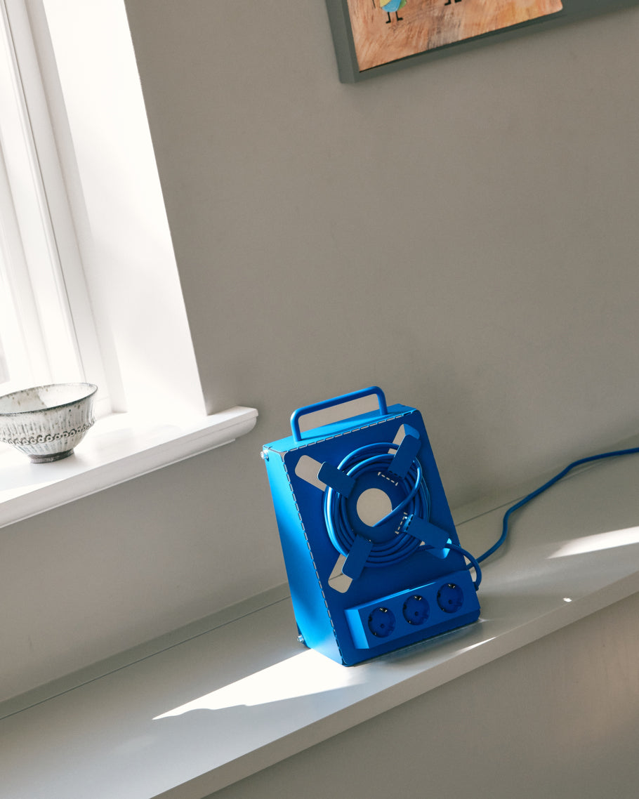 Ultra Marine Cable Stand on a sunlit windowsill ledge, angled view showing coiled cable, carry handle and three sockets