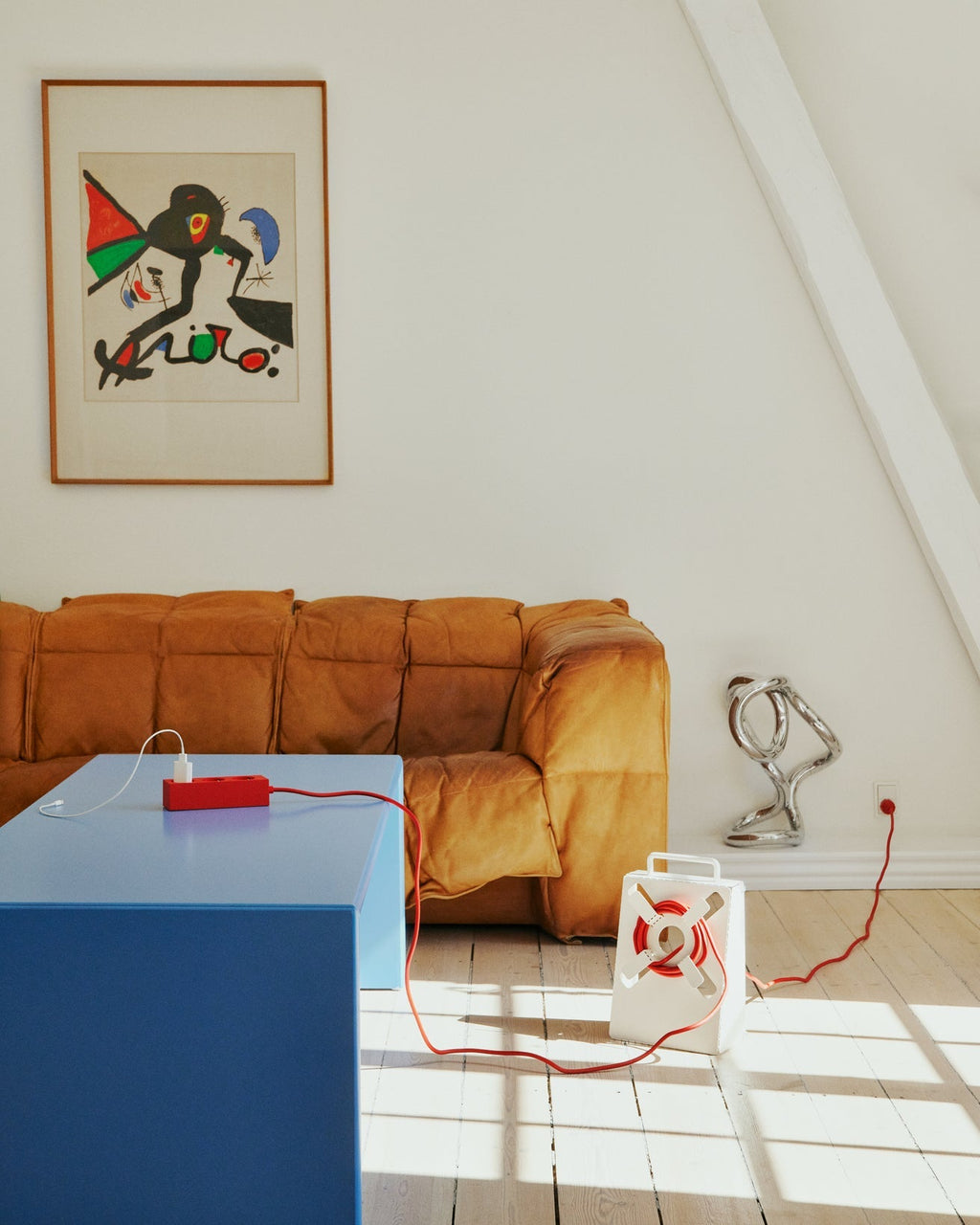 Pearl Cable Stand on sunlit wood floor, red cord unwinding to a blue table and outlet in a bright living room
