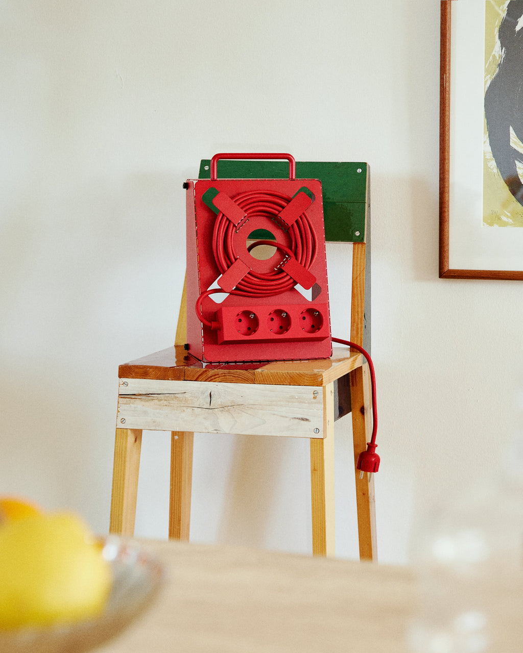 Fire Red Cable Stand with coiled cord and three sockets resting on a wooden chair in a sunlit room, plug hanging down