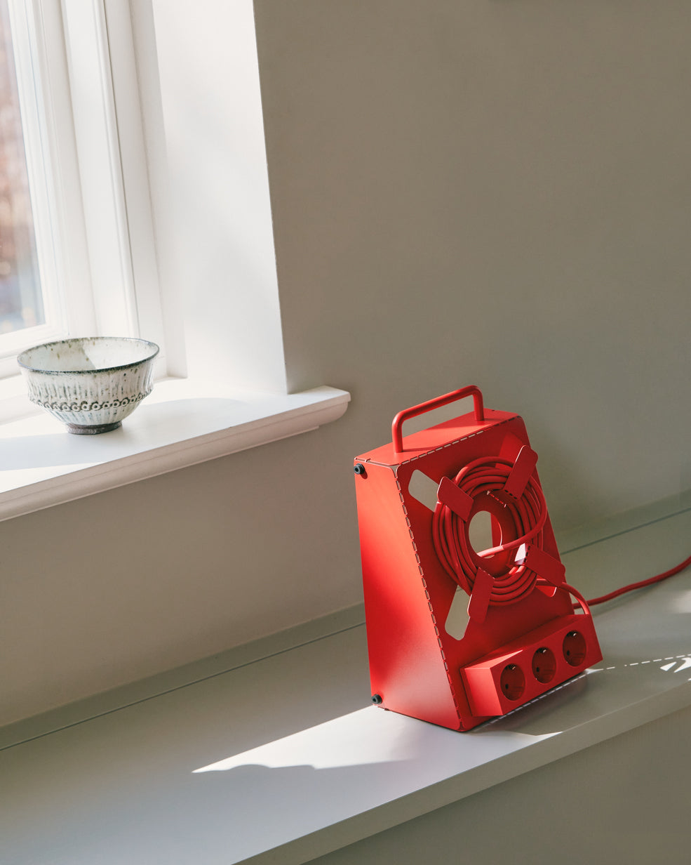 Fire Red Cable Stand with coiled cord and three sockets on a sunlit windowsill ledge beside a ceramic bowl