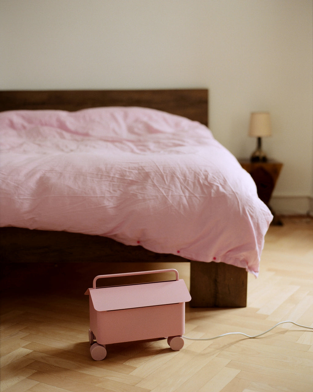Cable Villa in Bubble Gum on wheels beside a pink-linen bed, lid closed with white cable trailing across parquet