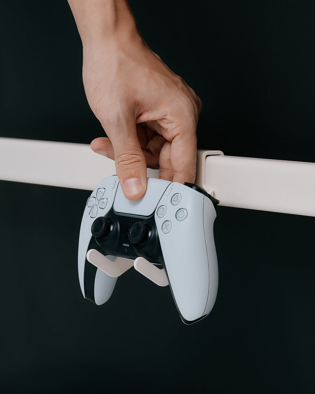 Hand placing a white PS5 controller onto the Controller Mount in Charcoal fixed to a wall rail against a dark backdrop