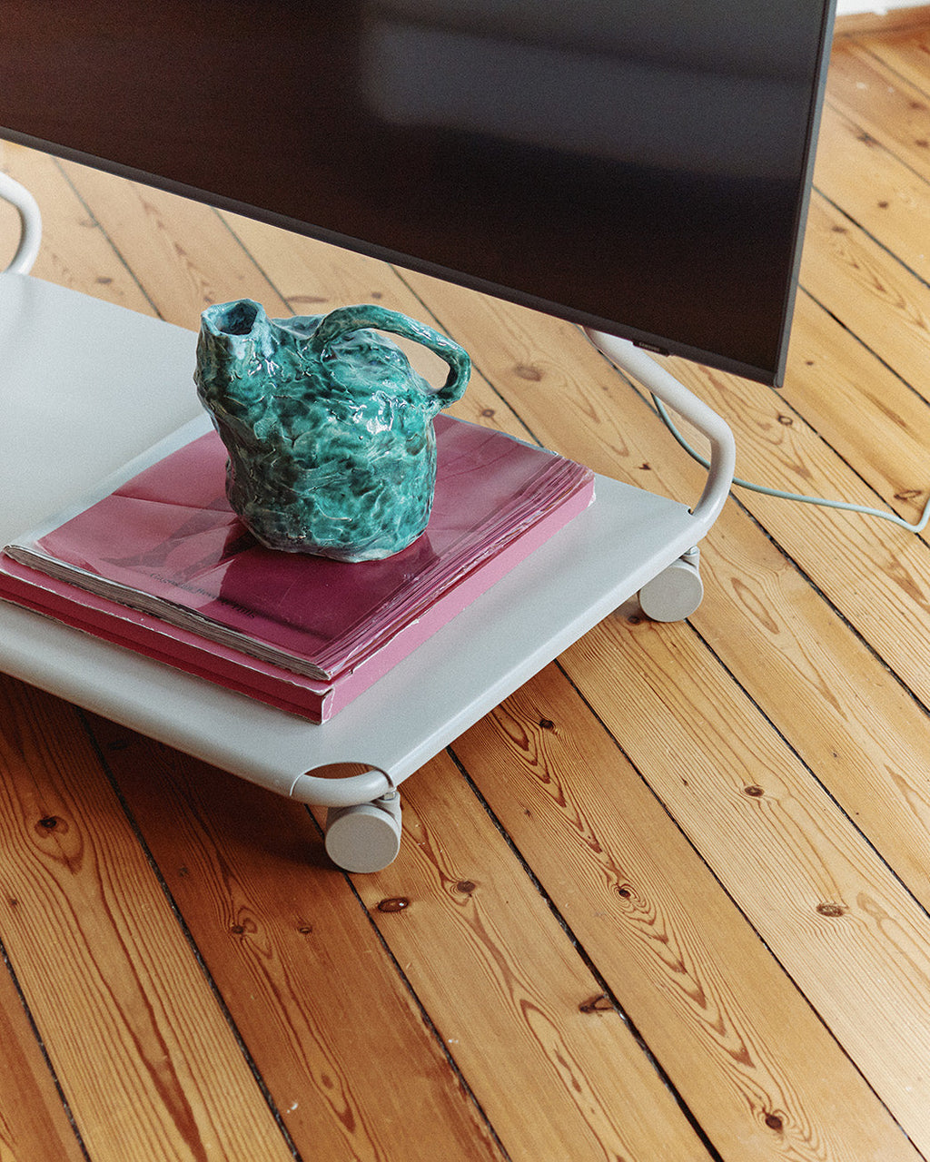 Disco stand in Oat on wheels holding pink books and a green ceramic vase beneath a TV on a wood floor