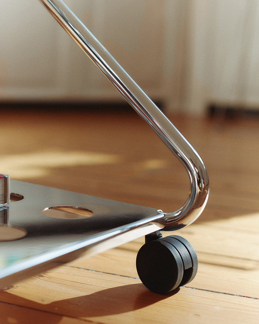 Close-up of Disco stand in Chrome on wood flooring, showing curved tubular leg and black caster wheel in sunlight