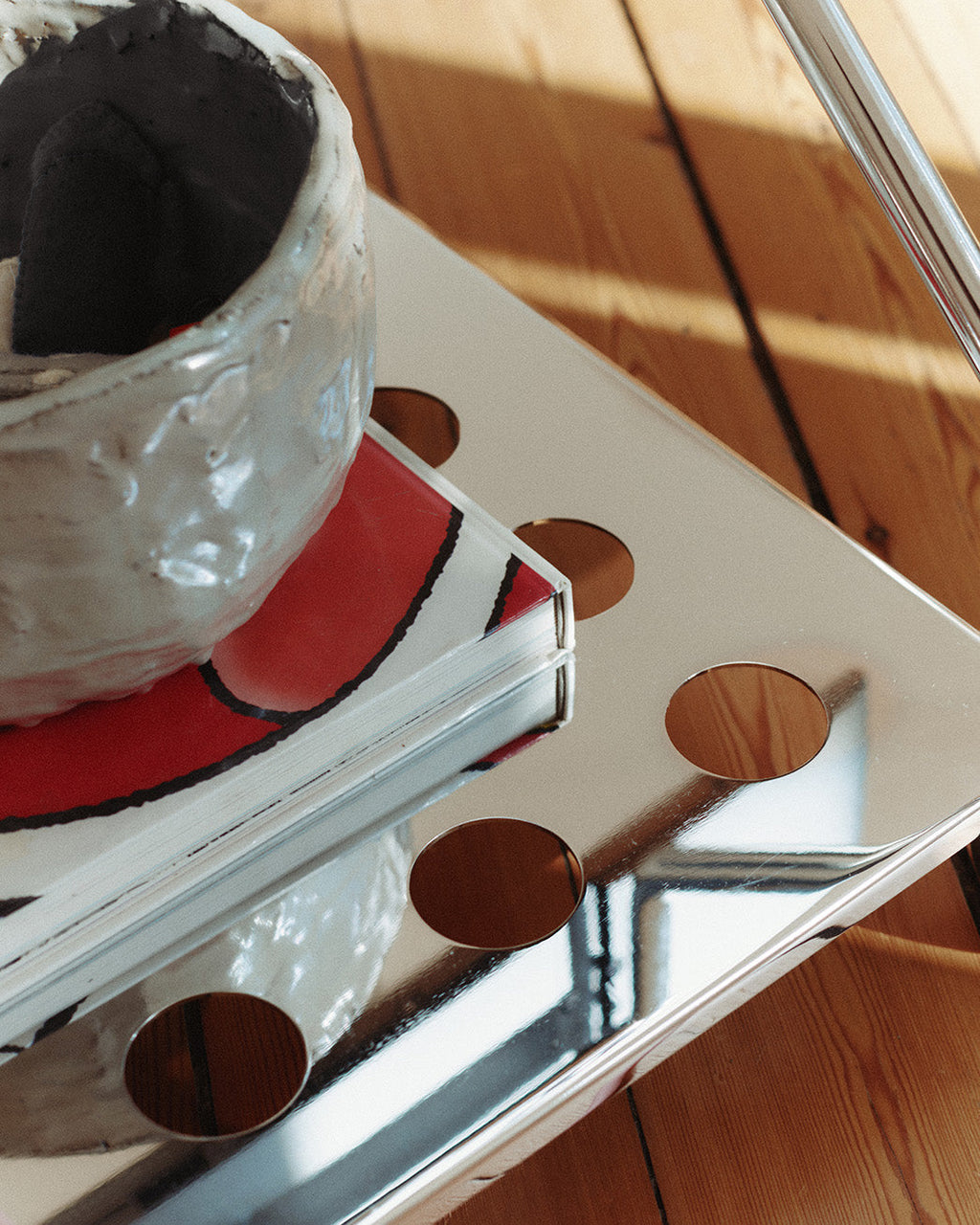 Close-up of the Disco stand in chrome, perforated top shelf holding books and a ceramic vase on wood floor