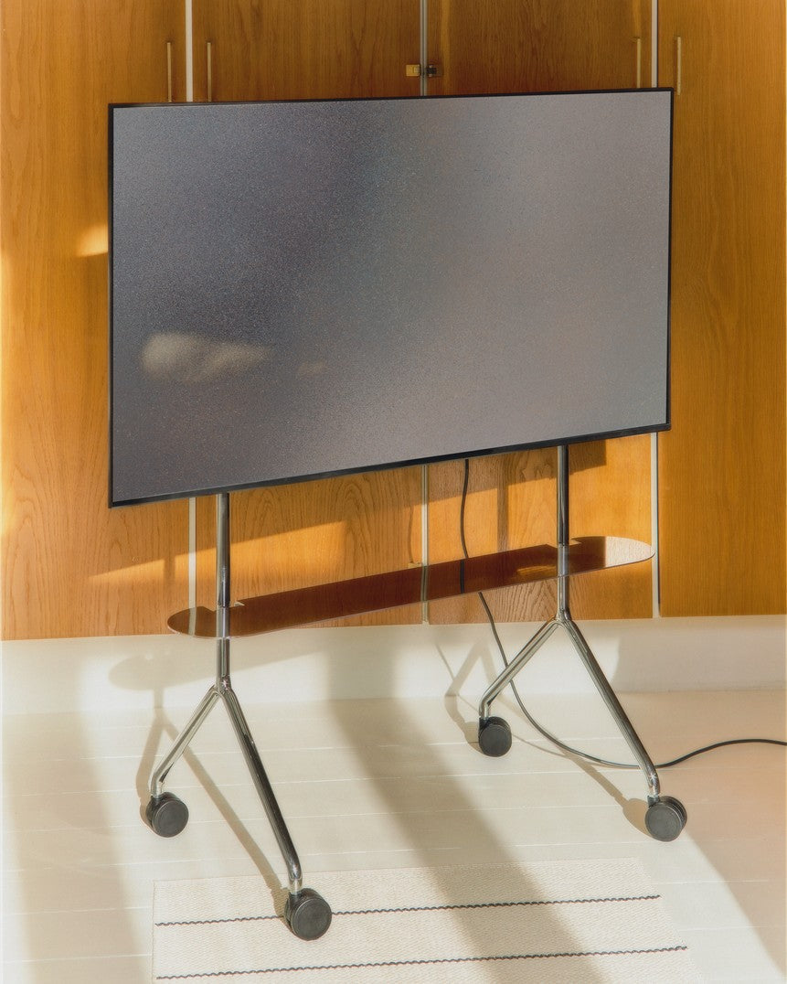 Moon Light TV Stand in chrome holding a large flat-screen in a sunlit wood-paneled room, front view on rolling casters