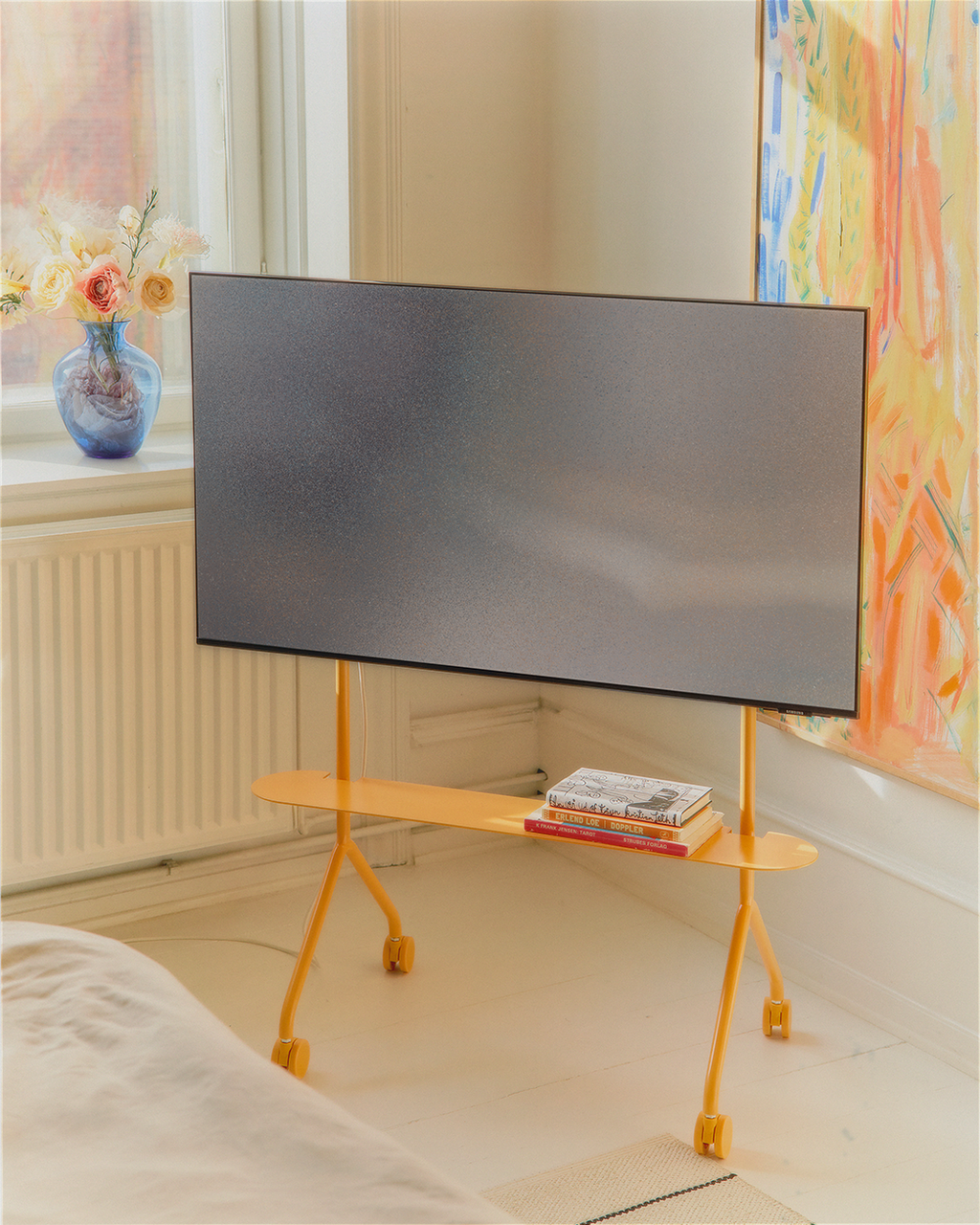 Moon Light Shelf in Apricot on wheels holding books beneath a mounted TV in a bright, art-filled bedroom corner