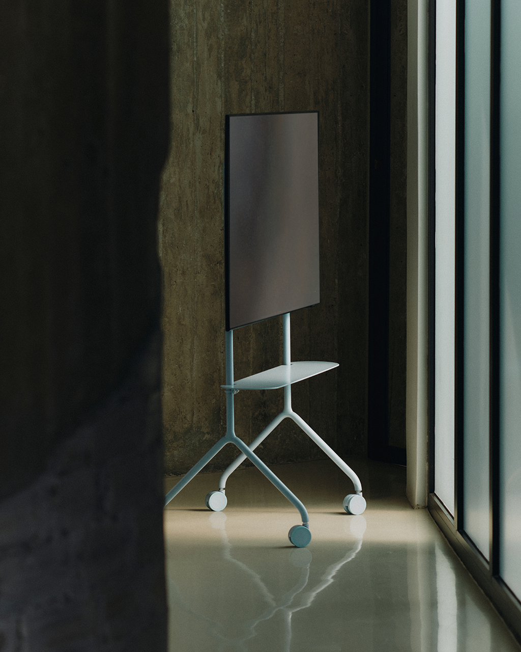 Moon Light Shelf in Powder Blue on wheels holding a mounted screen in a sunlit concrete room, angled side view