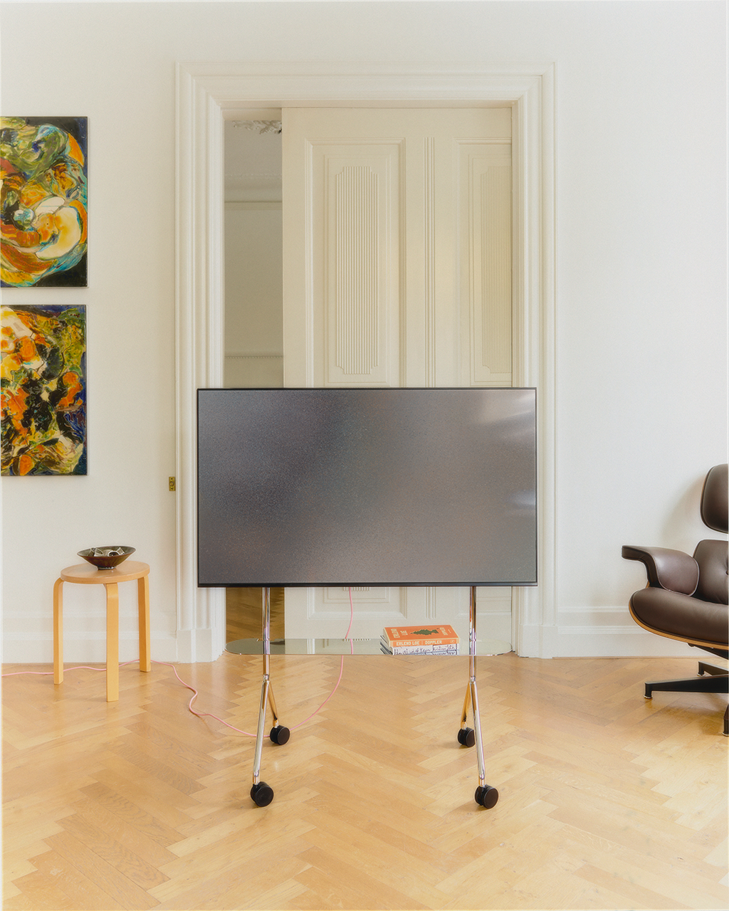 Moon Light Shelf in Chrome holding a large flat-screen TV on wheels in a bright living room with herringbone floors