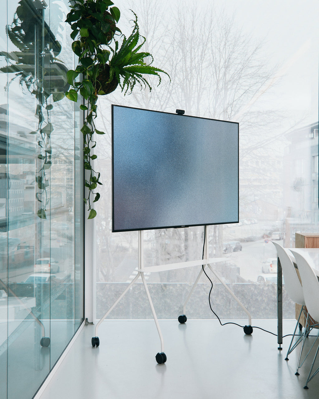 Moon PRO TV stand in Pearl on casters holding a large TV in a bright office by floor-to-ceiling windows and plants