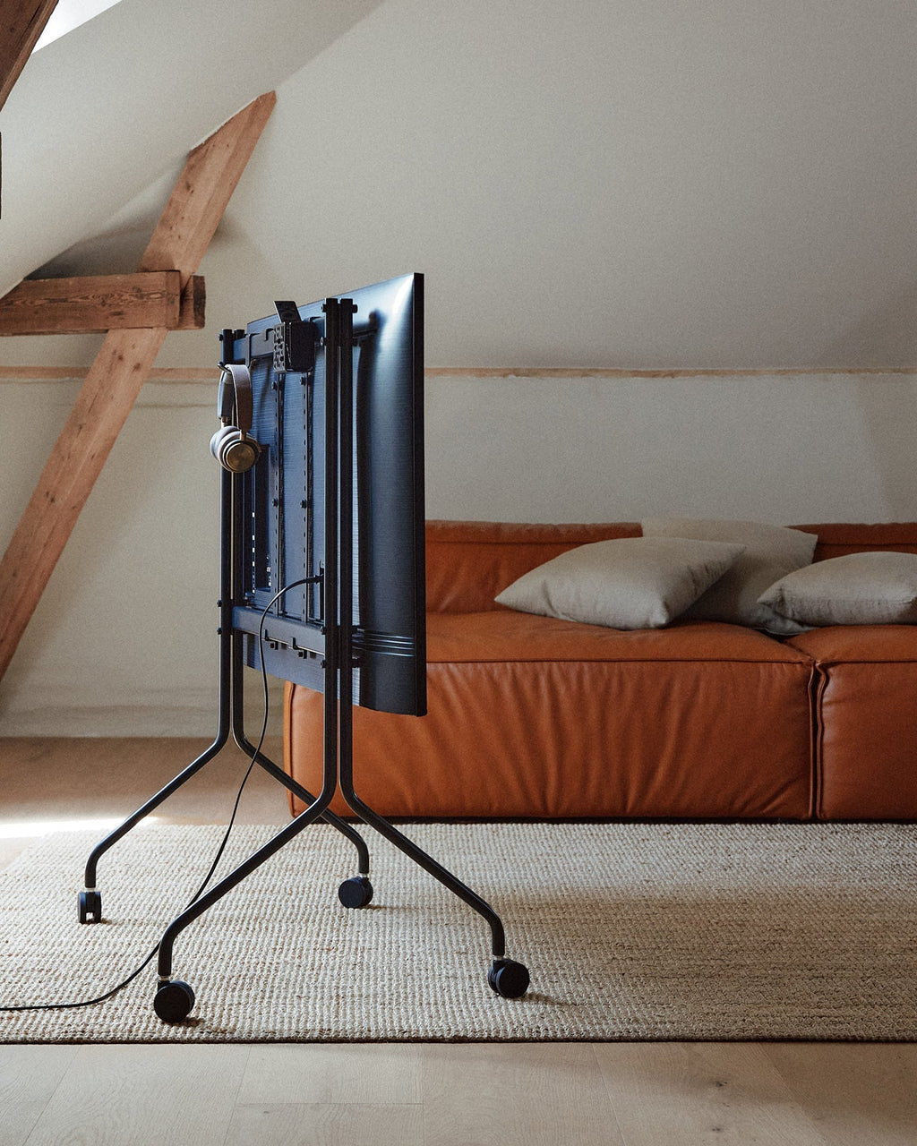 Rear view of Moon in Charcoal rolling TV stand with wheels in an attic lounge, headphones hung beside a tan leather sofa