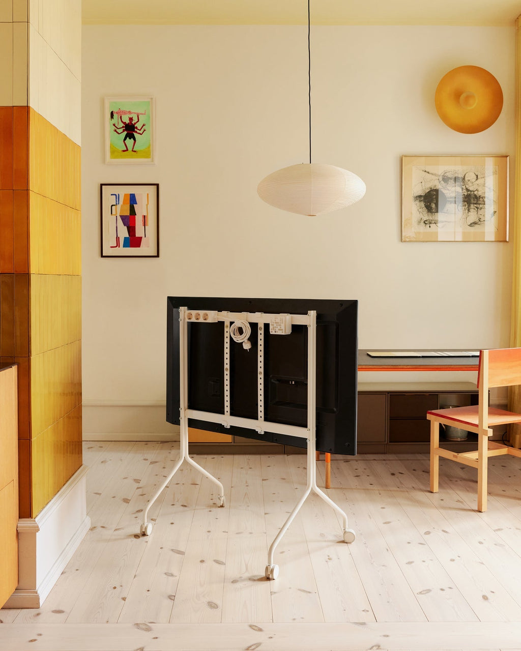 Rear view of Moon in Pearl stand with wheels holding a TV in a bright art-filled room with wood floors