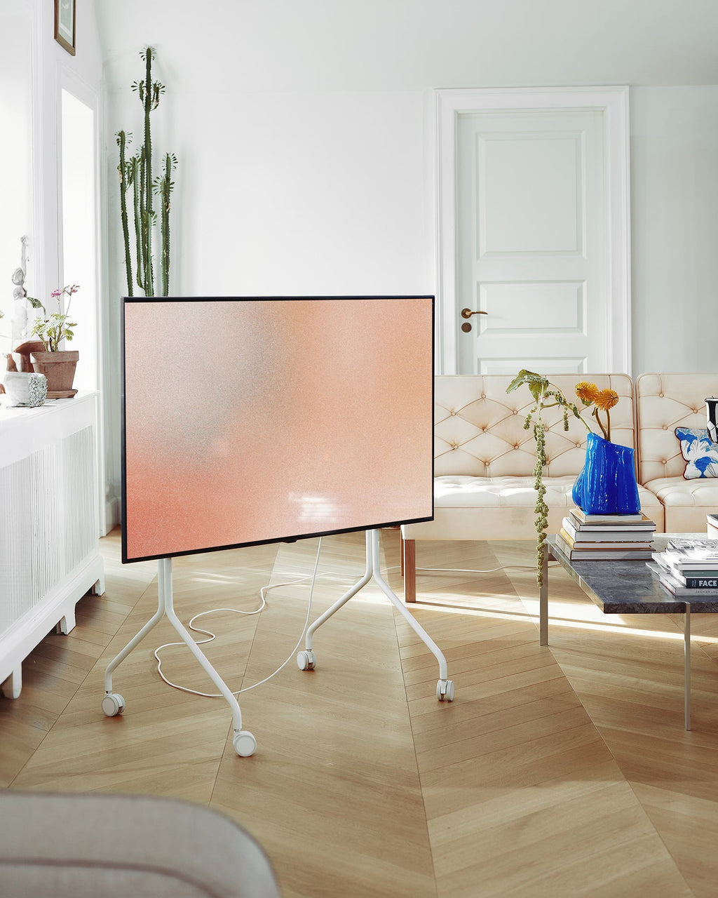 Moon in Pearl rolling TV stand holding a flatscreen in a bright living room with herringbone floors and tufted sofa