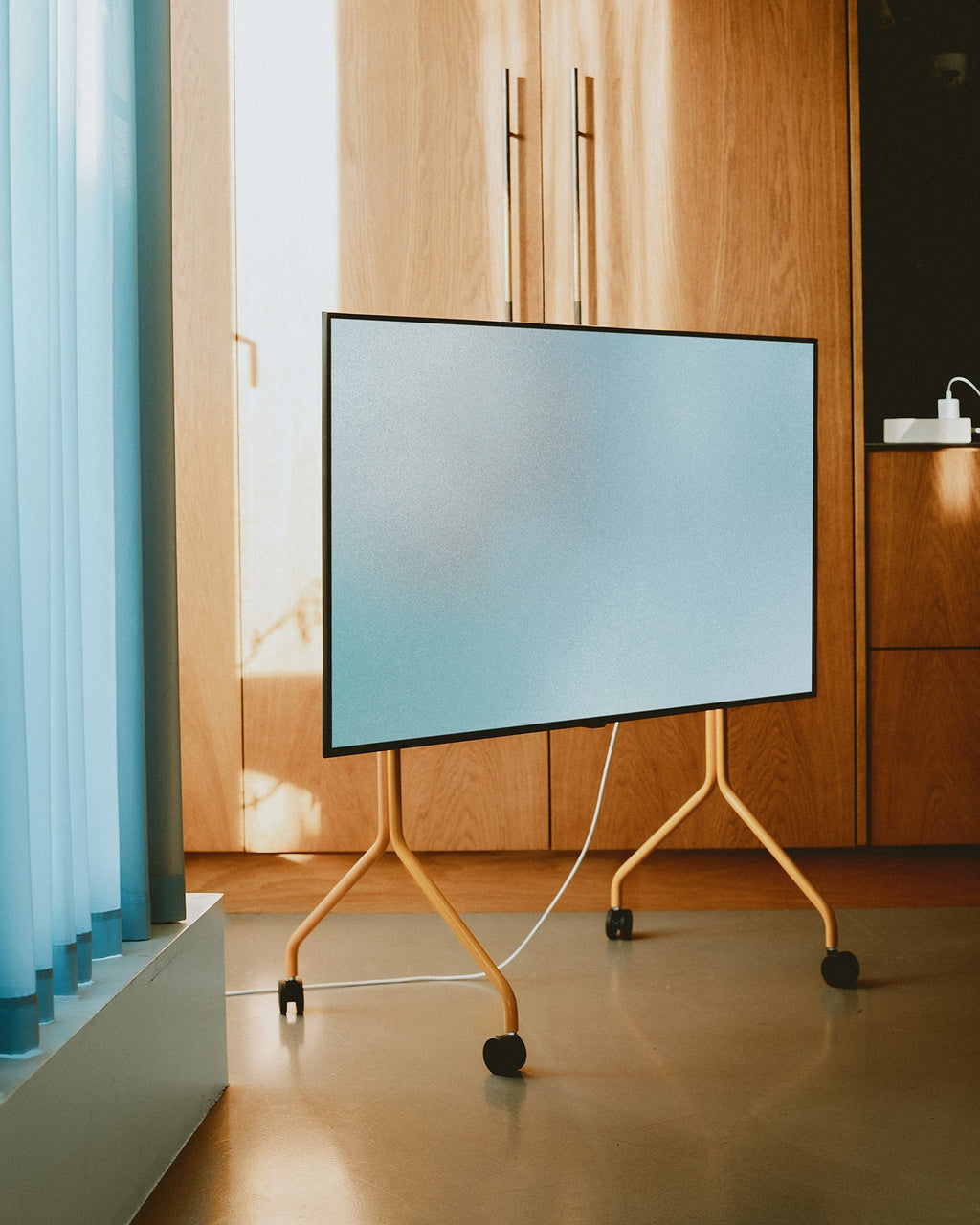 Moon TV stand in Apricot on wheels holding a large screen in a sunlit room with wood cabinetry and blue curtains