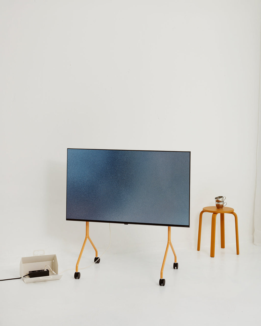 Moon TV stand in Apricot on wheels holding a large screen in a bright studio, beside an orange stool with cups