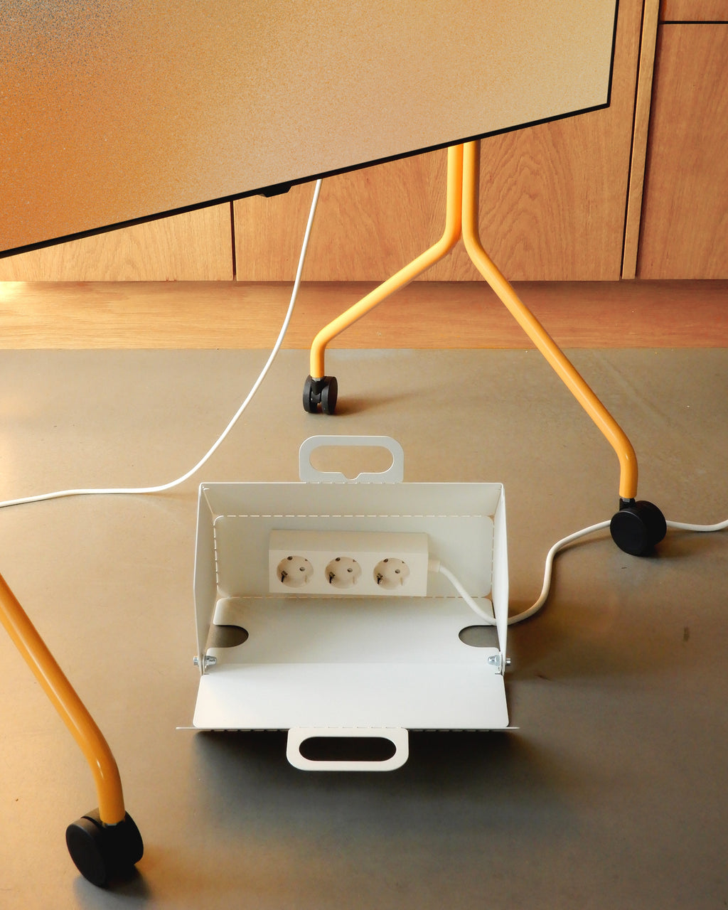 Close-up of Moon in Apricot stand's wheels and white cable box holding a power strip on a floor by wood cabinets