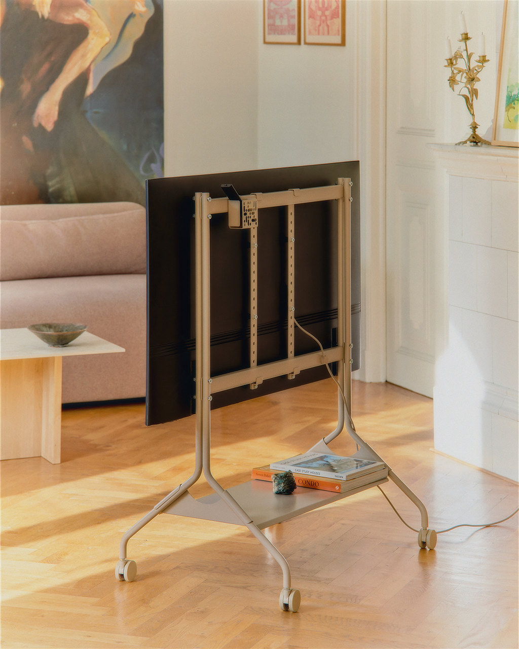 Moon TV stand in Oat holding a mounted TV, rear view in a sunlit living room with books on its shelf and rolling wheels