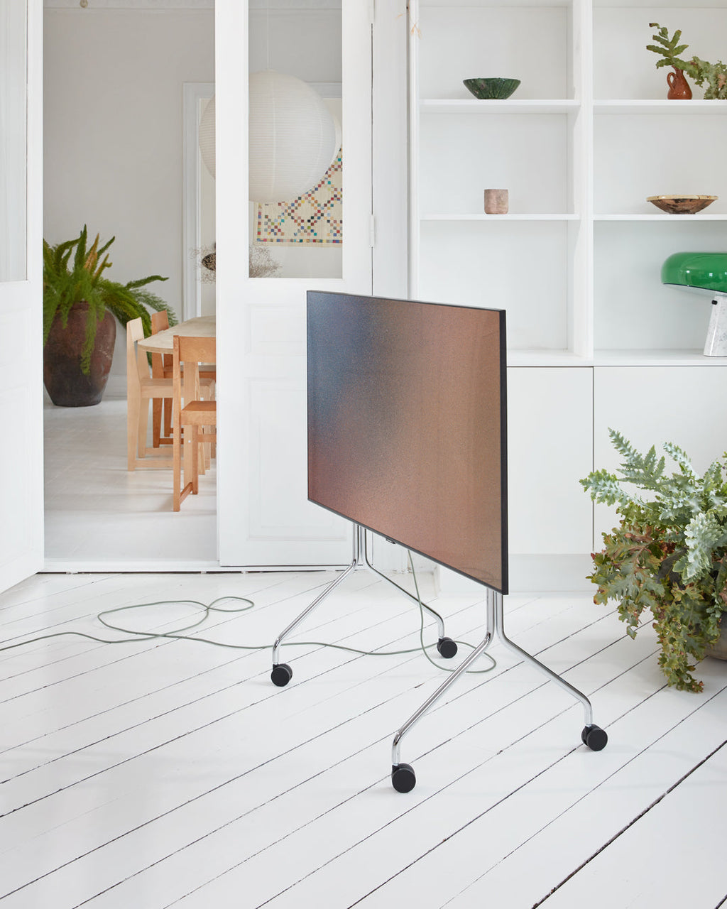 Moon in Chrome stand with wheels holding a folded-down TV in a bright white room with plants and shelving