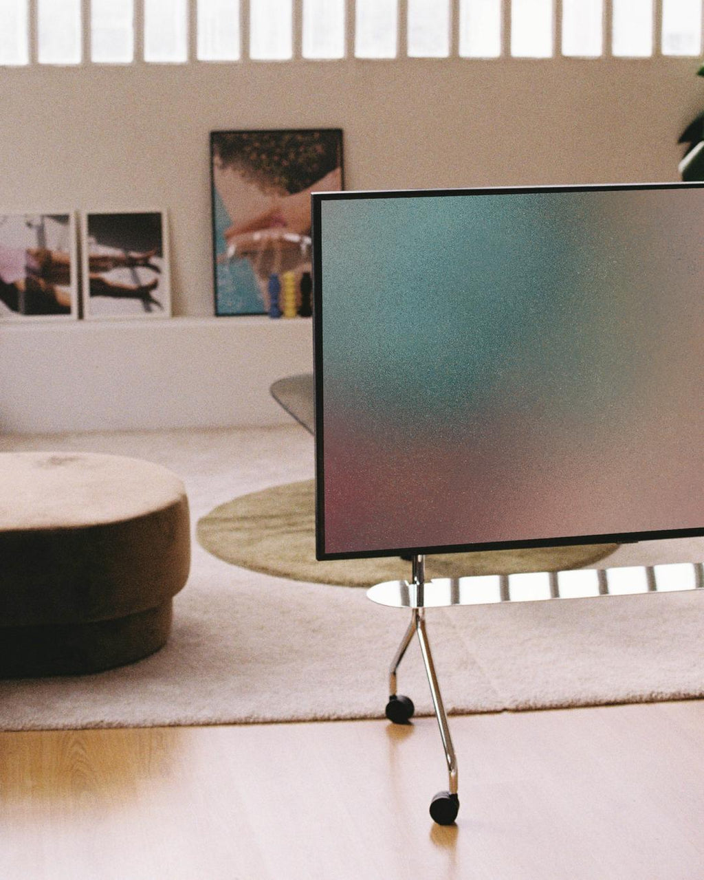 Moon chrome TV stand with wheels holding a flatscreen in a sunlit living room with rug, ottoman and framed art