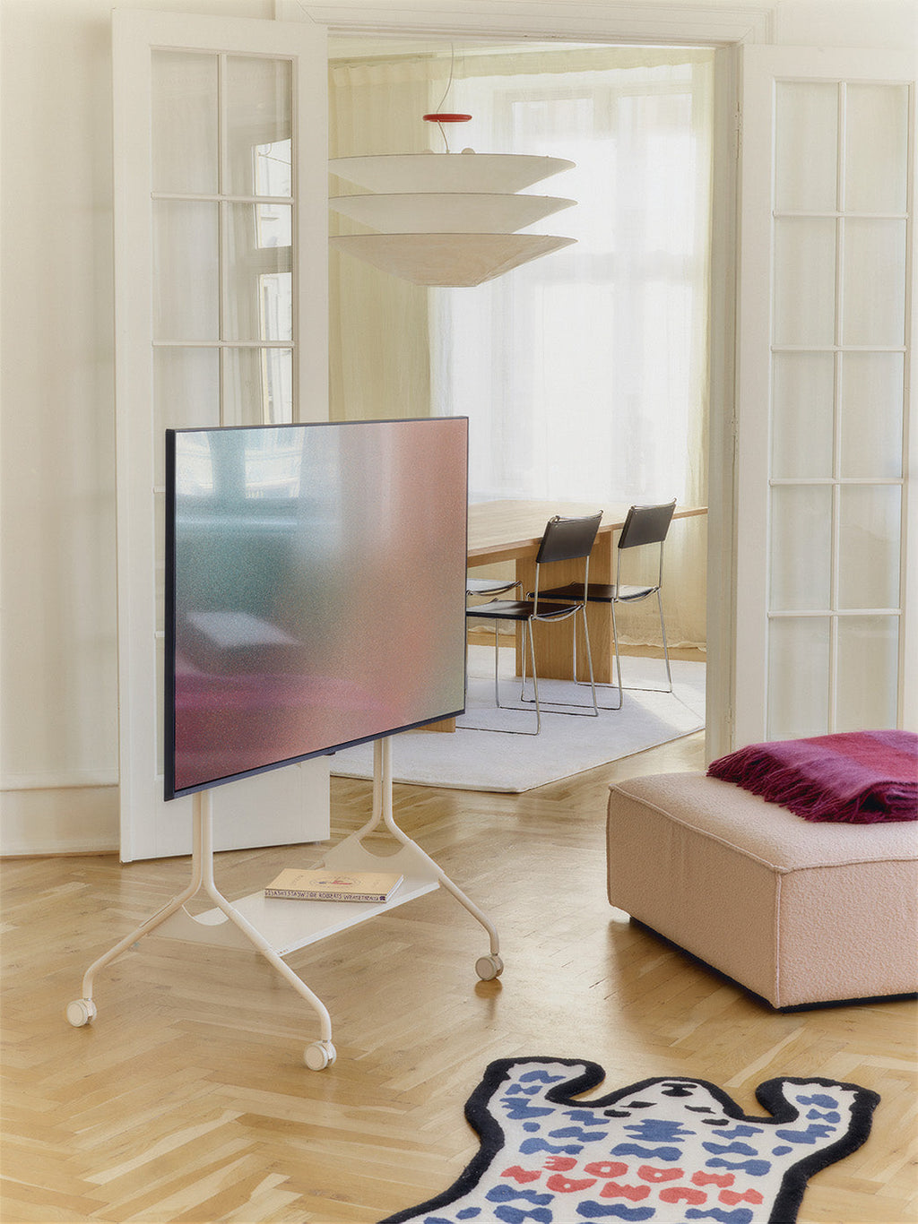 Moon Tall stand in Pearl on wheels holding a TV in a sunlit living room, with books on its lower shelf