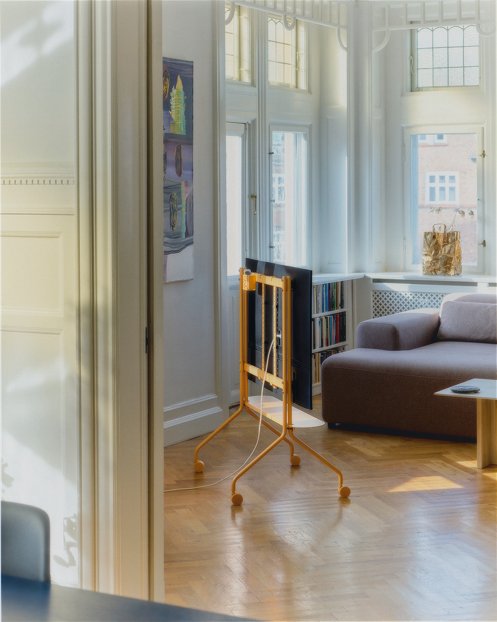 Apricot Wheels on a rolling TV stand holding a flat screen sideways in a sunlit living room with parquet floors