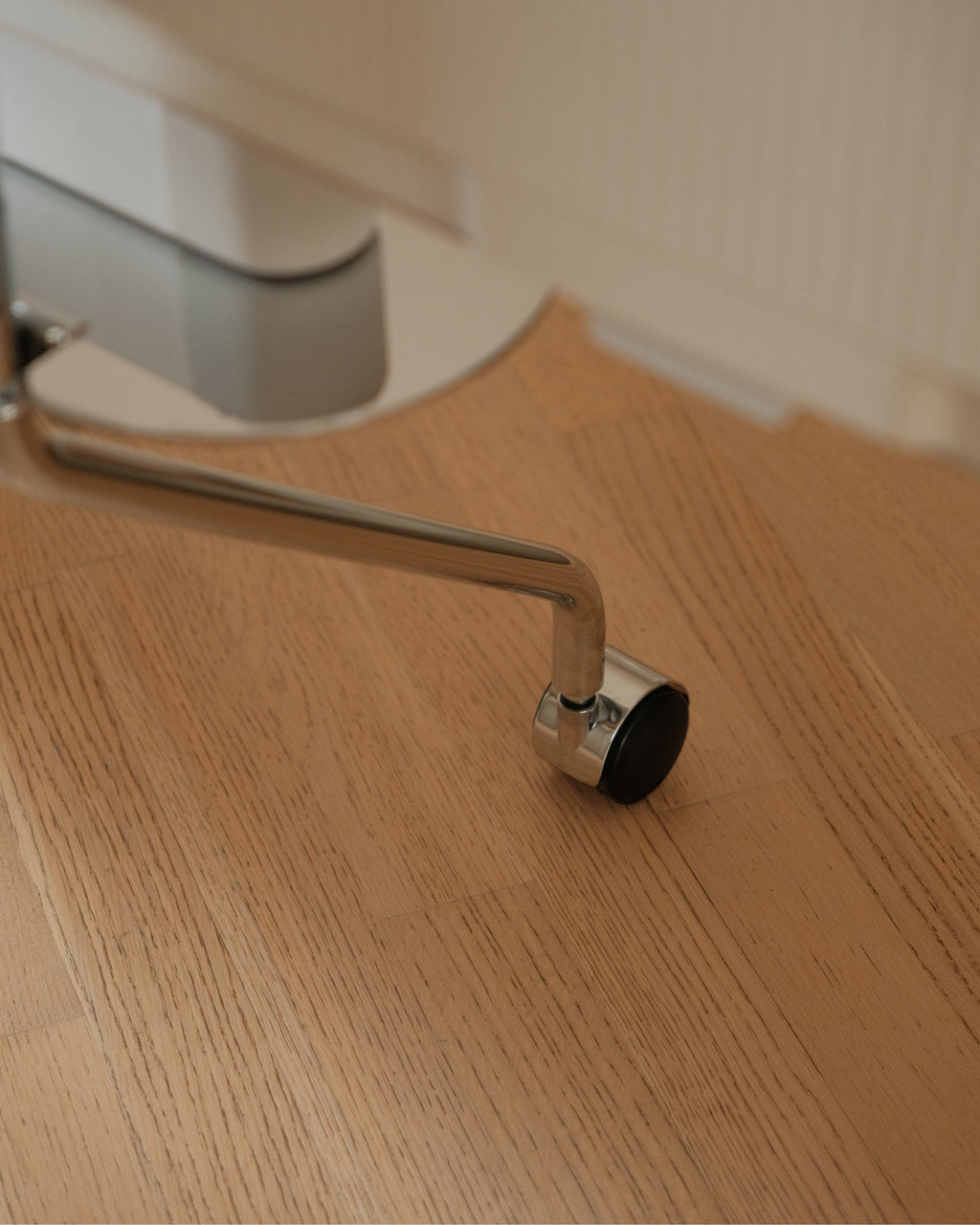 Close-up of a chrome Wheels caster on a light oak floor, showing the polished metal arm and black rolling wheel