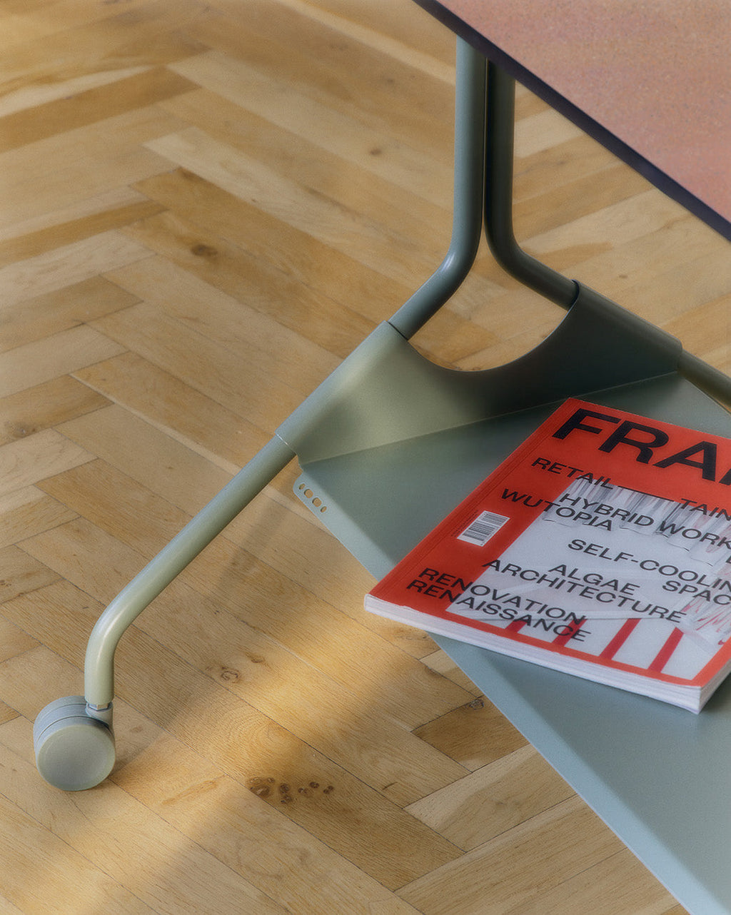 Moon Tray Shelf in Mossy Green under a desk on herringbone wood floor, holding a magazine, wheeled leg in focus