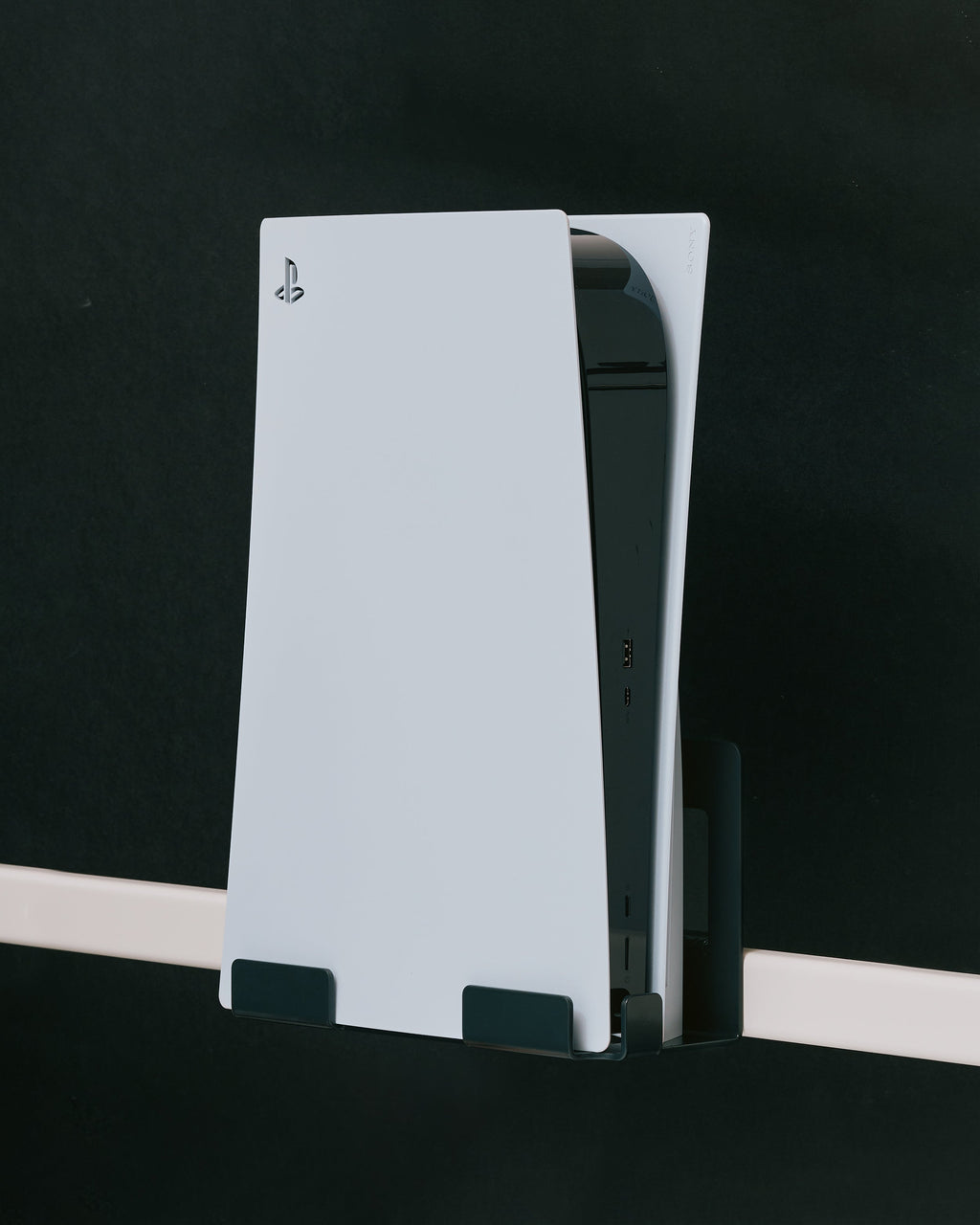 Pearl PS5 Mount holding a white PlayStation 5 vertically on a dark wall, angled side view showing USB ports
