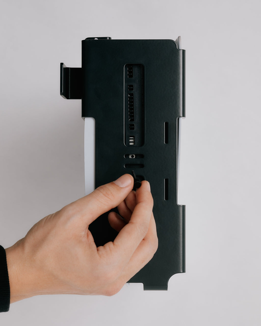 Hand inserting a mounting screw into the Pearl PS5 Mount held against a plain studio-grey wall, close rear view