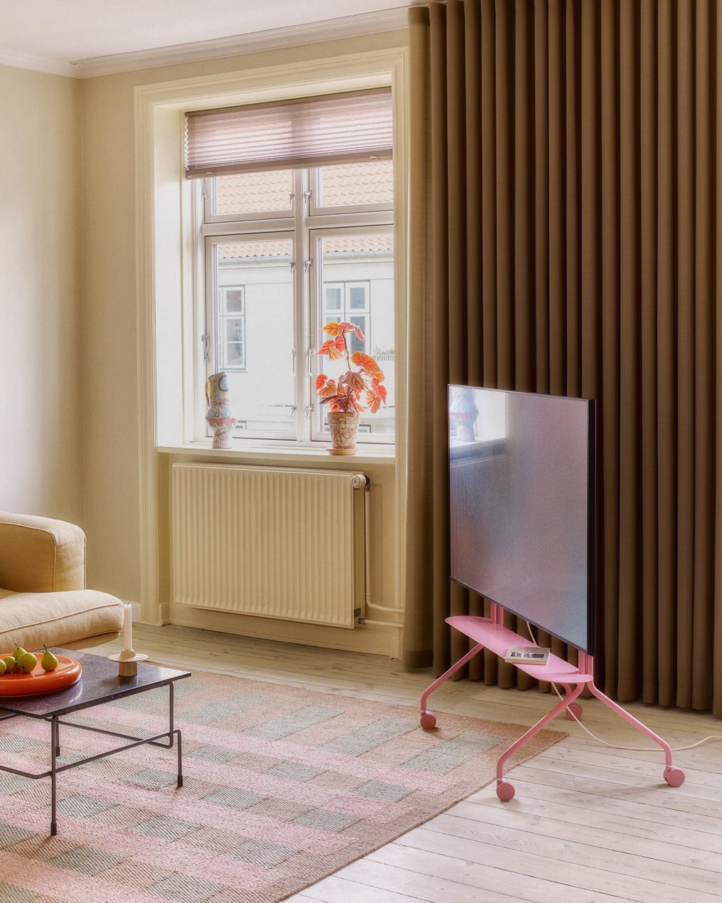 Moon Shelf in Bubble Gum holding a TV on rolling wheels in a sunlit living room with curtains and pastel rug